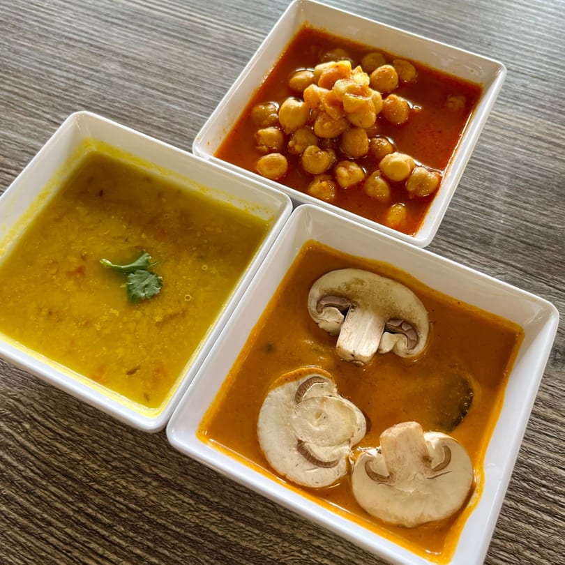 Three square white bowls of Indian cuisine: chickpea curry, yellow lentil dal, and mushroom curry.